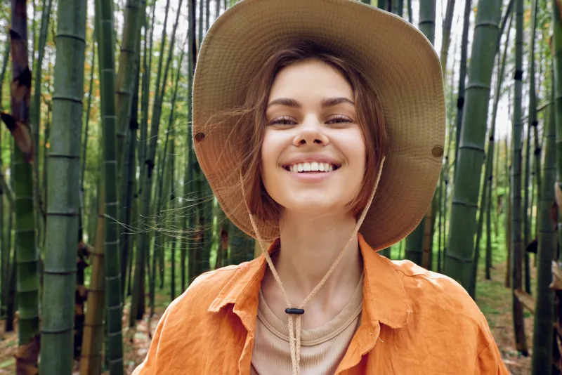 Woman smiling portrait in straw hat among bamboo forest, outdoor nature scene with casual orange shirt, cheerful traveler enjoying tropical greenery and peaceful relaxed mood.