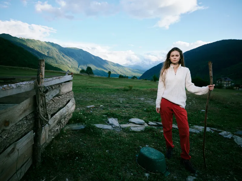 Woman standing in a green countryside valley with distant mountains, holding a wooden staff, wearing a white blouse and red pants beside a weathered fence in open fields.