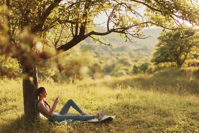 Young woman relaxing under a tree in a sunlit meadow, enjoying her phone amidst the serene natural landscape, representing tranquility and leisure in nature