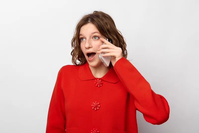 Surprised young woman in red sweater talks on smartphone with open mouth and expressive eyes. Isolated on white background, concept of communication, emotion, and technology.