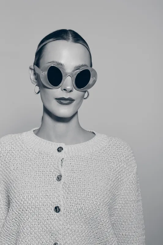Portrait of a fair skinned Caucasian woman wearing round sunglasses and a knitted cardigan isolated on light background. Fashion and style concept.
