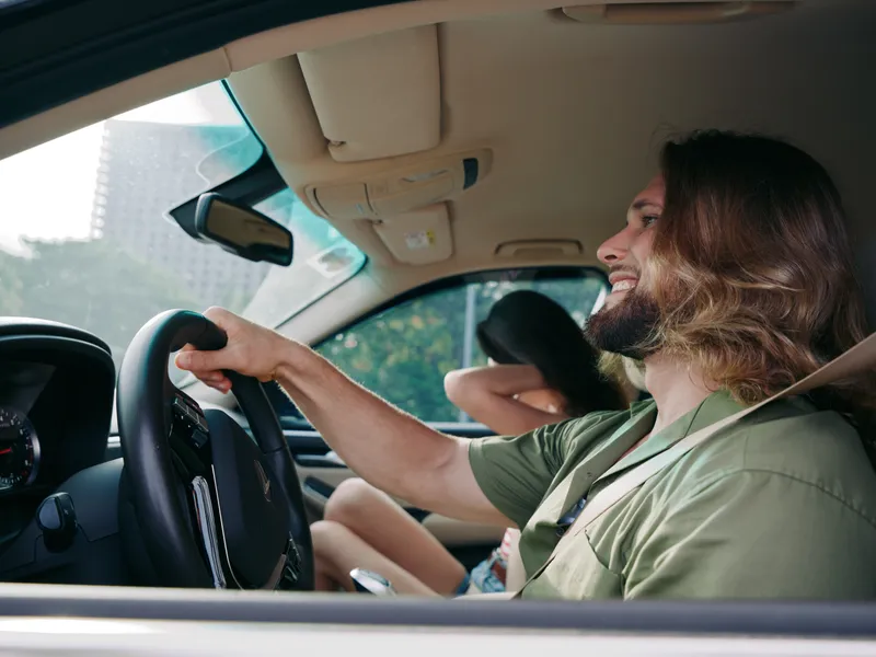 A smiling man drives a car with a laughing passenger in the back seat, capturing a candid moment of joy, travel, and casual road trip vibes under bright daylight.