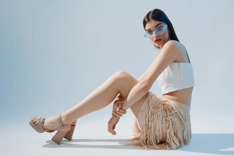 Young woman in fashionable sunglasses and a white top poses sitting on the floor against a light background. Concept for beauty, fashion, and style.