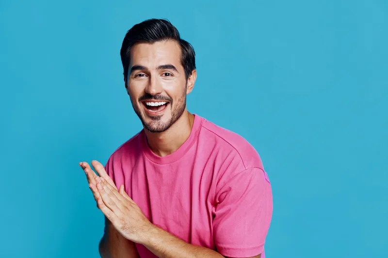 Happy smiling man in a bright pink shirt, looking at the camera with joy, hands together, isolated on a solid blue background. Lifestyle and positivity concept.