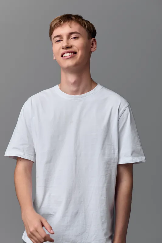 Smiling man in a white t shirt stands against a neutral gray backdrop, a casual studio shot conveying a relaxed, youthful style and clean fashion