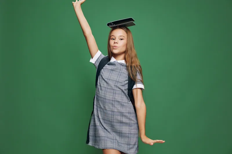 Girl student in school uniform posing with book on head and backpack on green background. Child with long hair standing and smiling for education and learning concept.
