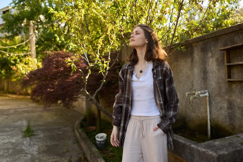 Young woman outdoors in a plaid shirt over a white tee stands in a sunlit garden, looking upward with a relaxed, curious expression as a gentle breeze moves her hair
