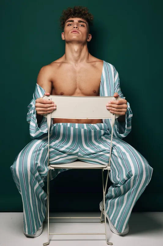 Young man with curly hair sitting on white chair backward wearing striped loose pants and open shirt showing bare chest against dark green background in studio.