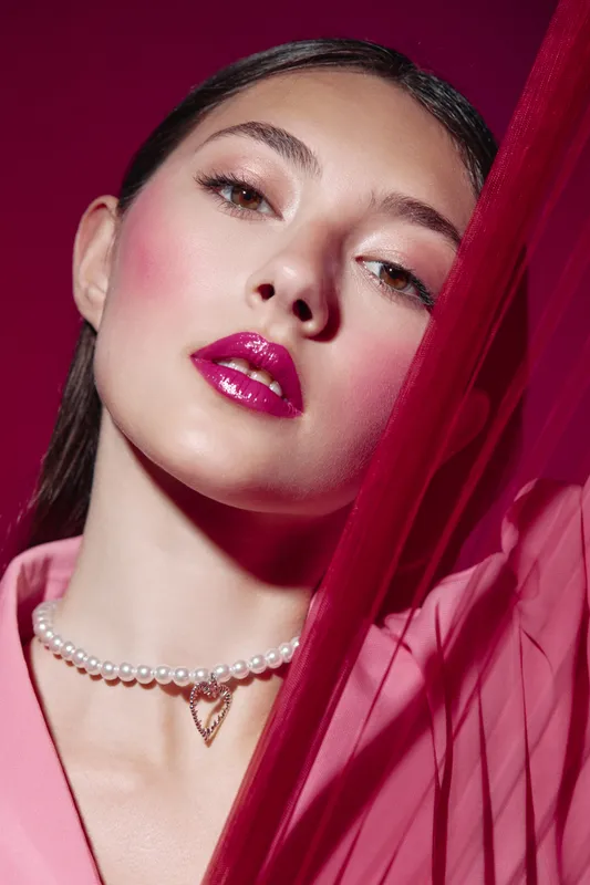 Close-up portrait of young woman with glossy pink lips and vibrant blush wearing pearl necklace and pink blouse, posing with red fabric. Concept of beauty, creative fashion, and makeup art.