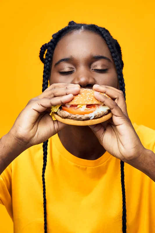 Young Black woman joyfully enjoying a delicious hamburger against a vibrant yellow background, embodying the concept of fast food and happiness