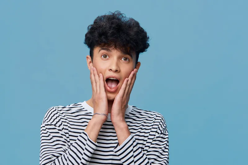 surprised man with curly hair and striped shirt expresses shock and amazement on blue background, showing emotion and youthful energy