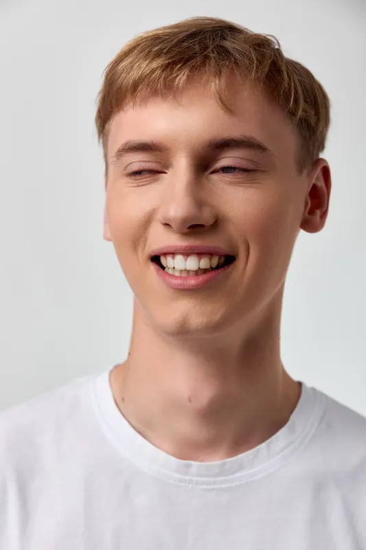 Smiling man with closed eyes wearing a white tshirt expressing happiness and a cheerful, relaxed mood in a soft studio setting, friendly and approachable vibe