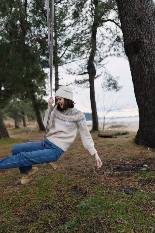 Outdoor swing scene with a woman in a knitted sweater and beanie enjoying a forest setting and autumn calm, capturing playful relaxation in nature.