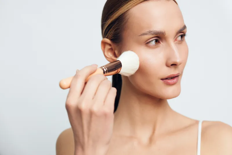 Young woman applying powder makeup with brush on clean skin, focused expression, natural light, minimalistic background, beauty routine, skincare, cosmetic application
