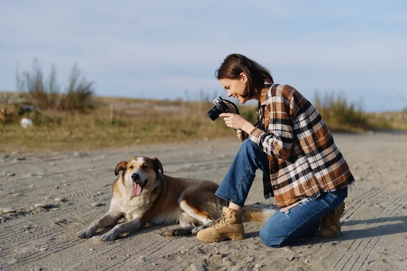 A photographer woman kneels on a sandy beach, camera ready, capturing a calm dog in a wide coastal landscape, highlighting light, texture and connection