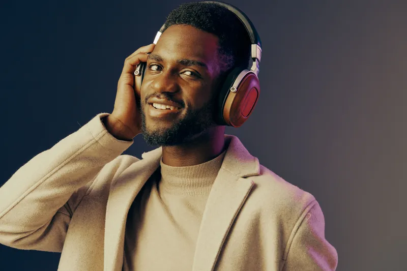 Young man enjoying music with headphones, smiling and stylishly dressed in a beige coat against a gradient background, representing relaxation and modern lifestyle