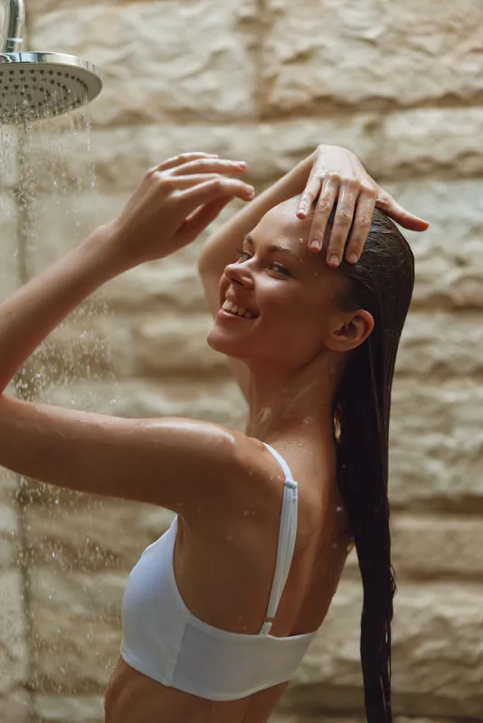smiling woman enjoys refreshing warm shower indoors, wet hair, casual white top, natural stone wall background, peaceful moment of self-care and relaxation