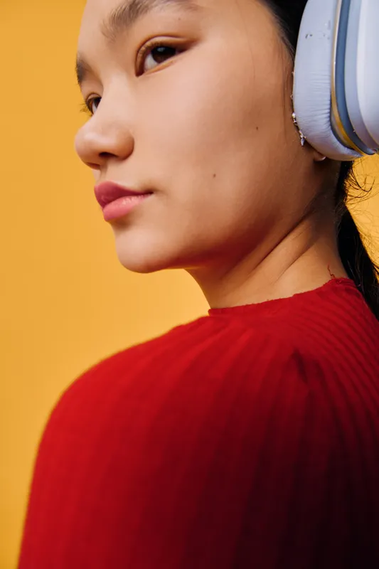 Portrait of young woman wearing headphones and red sweater with natural skin and serious expression indoors against yellow background with soft light and casual style.