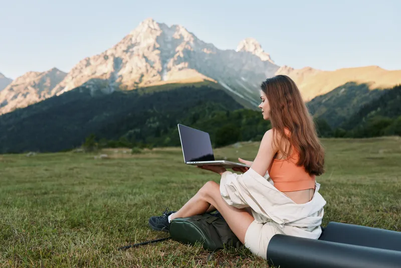 A focused young woman sits on a grassy hillside with a laptop, enjoying natural scenery. She works outdoors while towering mountains rise behind her, creating a serene and productive remote work vibe.