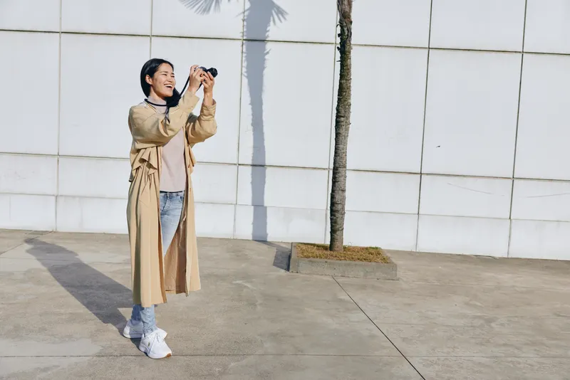 Photography of a woman in a beige trench coat outdoors, holding a camera against a white tiled wall with a palm tree, showcasing urban lifestyle, candid shooting, and creative image capture for stock