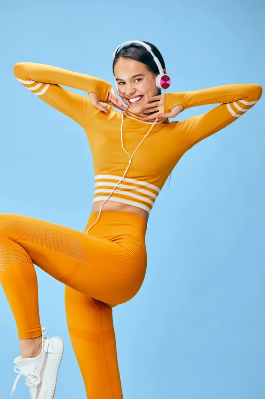 Woman in bright orange athletic outfit enjoying fitness and sport activity, posing with energetic movement, wearing headphones, demonstrating workout and exercise enthusiasm against a vibrant blue