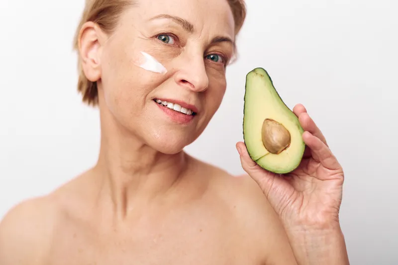 Mature woman with short hair holds half an avocado near her face with a smile. She has a small amount of face cream on her cheek, symbolizing natural skincare and healthy lifestyle.