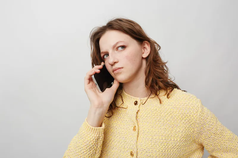 Young woman in yellow sweater talks on smartphone with serious expression, isolated on white background. Concept of communication, technology, and thoughtful conversation.