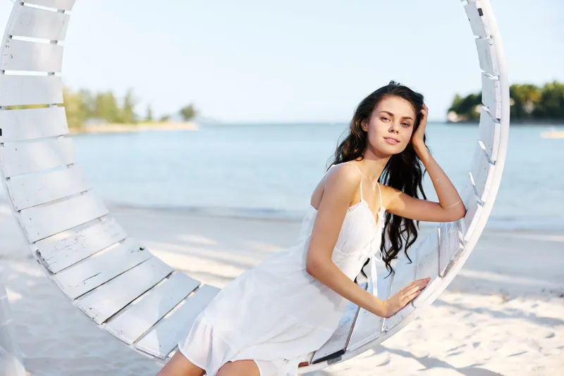A graceful woman in a white dress sits on a circular seaside swing, sunlit sand and gentle water frame a serene summer scene by the shore and calm horizon.