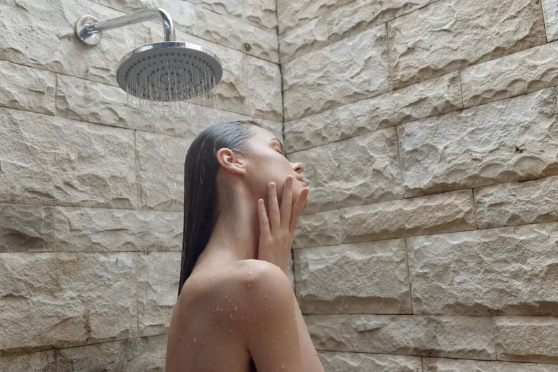 woman showering under rain showerhead with stone wall background, wet hair, relaxed skin care moment in natural light