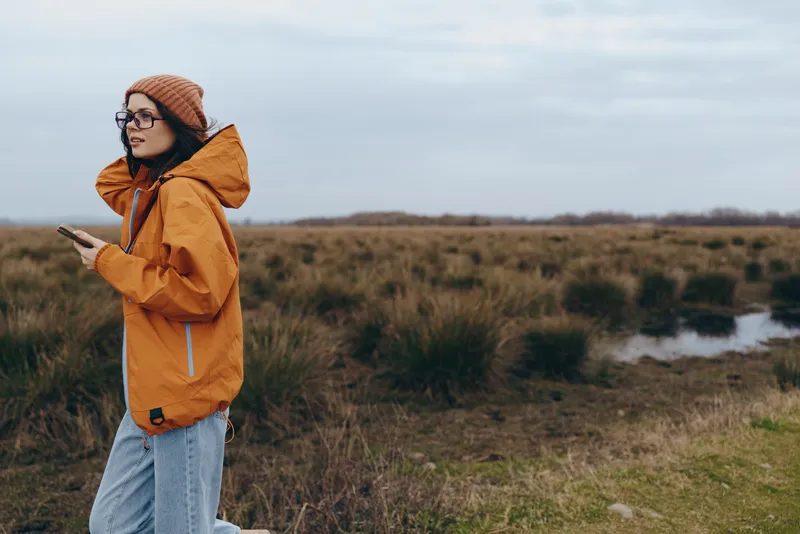 Woman lifestyle outdoors, standing in a field by a river, wearing orange jacket and knit hat, using a smartphone, calm travel mood, natural daylight, countryside