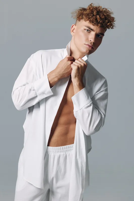 Young man with curly hair wearing white open shirt and pants posing against gray background, showing toned body and confident expression in studio lighting.