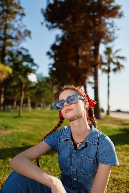 Lifestyle woman in modern boho-western denim street style with film color, wearing blue sunglasses and a red scarf, enjoying outdoor sunny park setting.