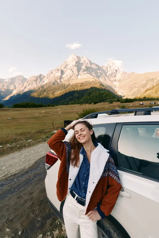 Woman leaning on SUV by roadside with mountain landscape, smiling and enjoying travel adventure in nature. Casual outfit with denim and jacket, relaxed pose and scenic outdoor view.