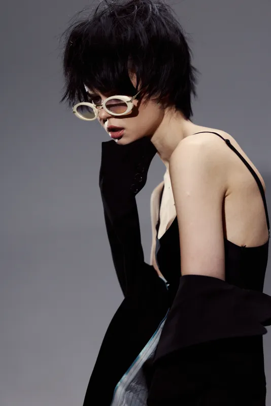 Stylish woman with short black hair and sunglasses posing in a black outfit against a gray background, expressing modern fashion and elegance.