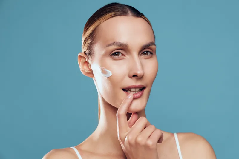 Woman skincare cream face beauty applying close up portrait spa, she applies moisturizer with fingertip on cheek, smooth glow, clean beauty routine, minimal blue background, natural light