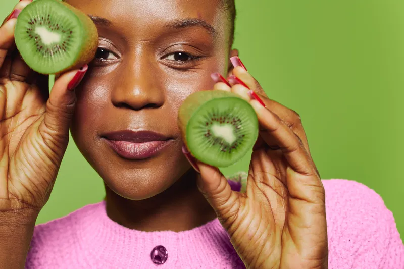 Closeup of a smiling black woman with dark skin holding two halves of kiwi fruit near her face on green background. Concept of healthy lifestyle, natural food, and fresh fruit.