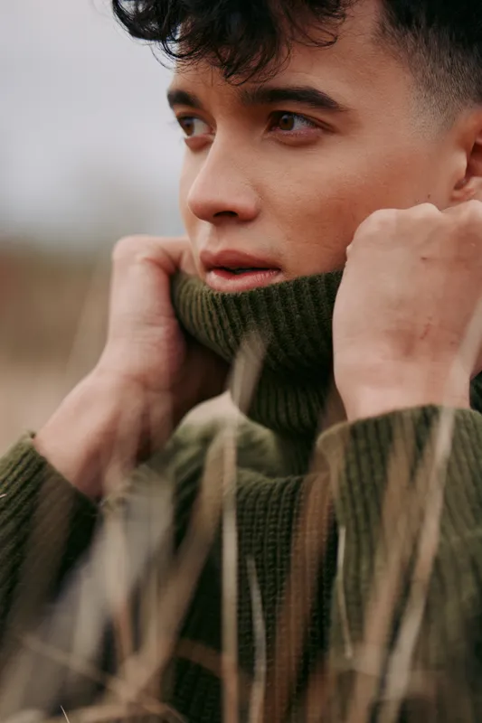 Close-up portrait of a young man pulling his turtleneck sweater up, conveying authentic presence and credible emotion with detailed facial texture and a natural, contemplative gaze.