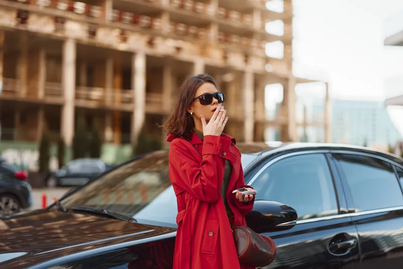 A stylish woman in a red trench coat stands beside a black car in an urban setting, wearing sunglasses and a confident casual vibe as she pauses near the vehicle