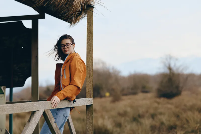 Woman lifestyle outdoors, seated by wooden fence, wearing orange jacket and glasses, autumn wind lifting hair, calm countryside field background, soft natural light, candid portrait, warm mood