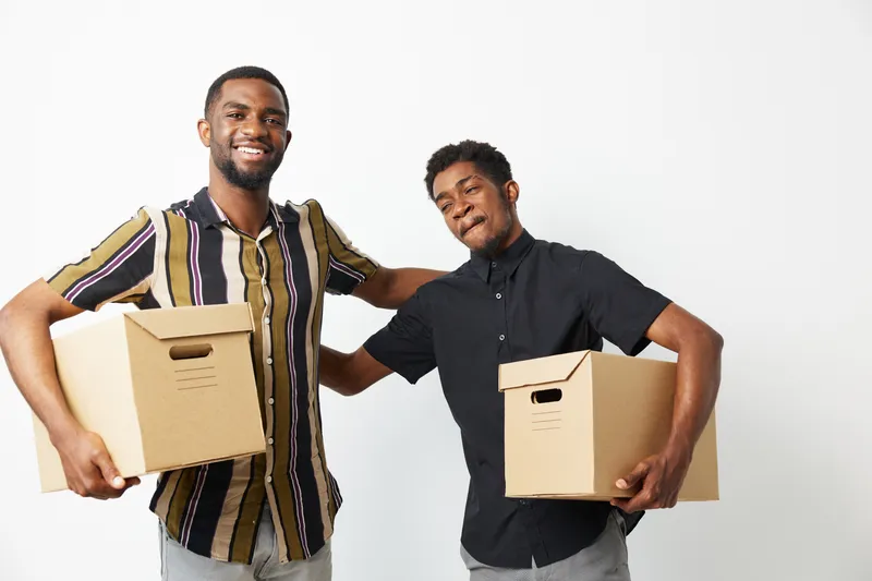 Two smiling African American men are standing close together, joyfully holding cardboard boxes. They express teamwork and happiness in a bright and cheerful environment.