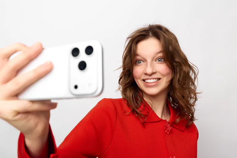 Smiling woman with curly hair wearing red sweater takes selfie with white smartphone on white background. Concept of social media, communication, and modern technology usage.