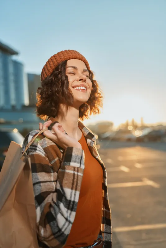 Woman with a smile and shopping bag at golden hour in an urban parking lot, candid autumn portrait with authenticity and mindful living, cozy beanie and plaid jacket.