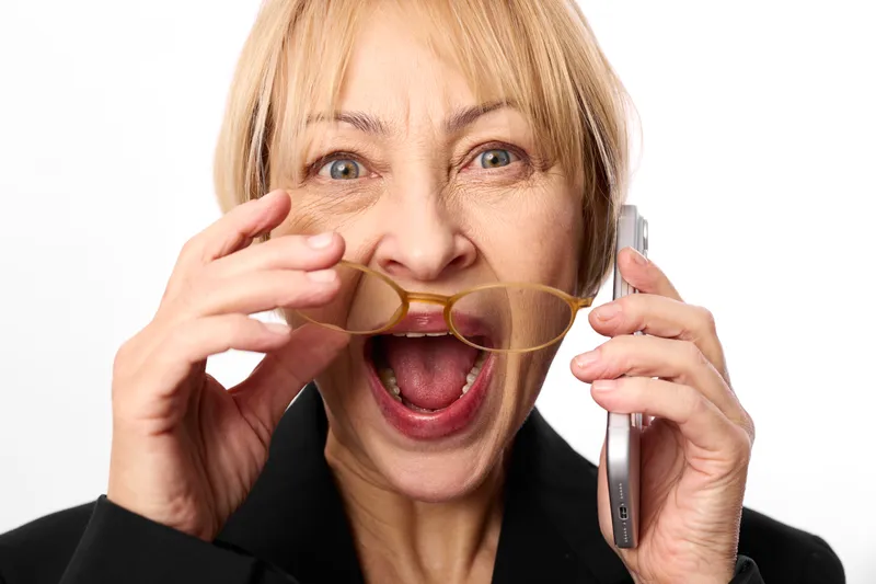 Mature blonde woman shouting into a megaphone, excited expression, holding cardboard megaphone, announcement concept, isolated on white studio background.