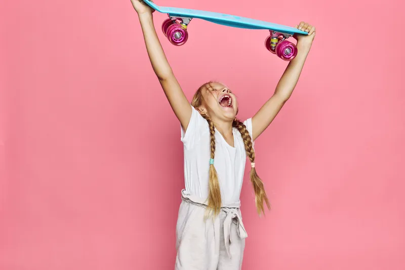 Excited girl with long braided hair lifts a skateboard above her head while shouting. She wears casual white t-shirt and light pants on a solid pink background. Joy and energy concept.