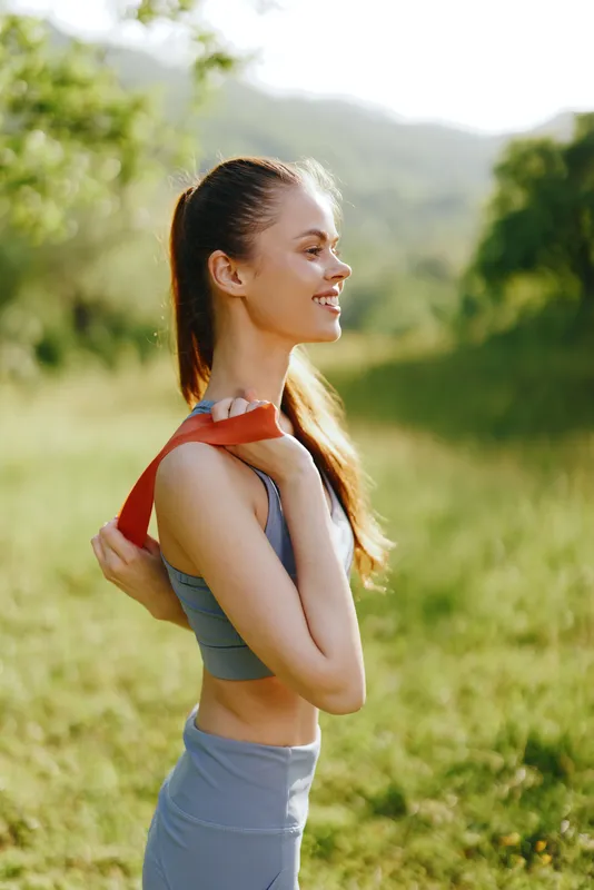 Young athletic woman smiling while exercising outdoors with resistance band, wearing sports clothing, showcasing fitness and healthy lifestyle in nature