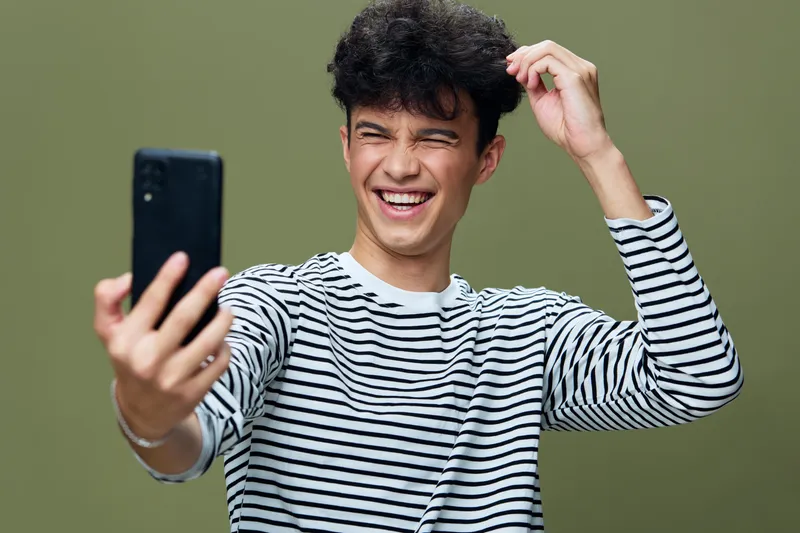 young man taking selfie in striped shirt smiling while scratching head with curly hair and olive green background, casual and joyful mood captured