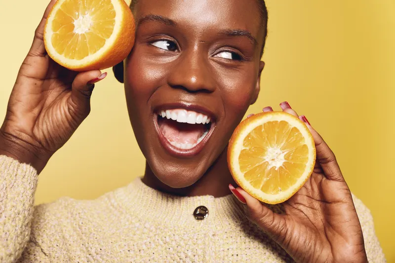 Happy African woman with dark skin holds two orange halves near face smiling widely. Bright yellow background and cozy beige sweater create warm, fresh, and joyful concept of health and fruit.