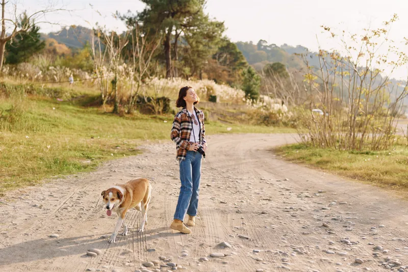 Woman walking a dog along a gravel path in a sunny countryside setting, wearing casual clothes, enjoying outdoor leisure and nature with open fields and trees in the distance.