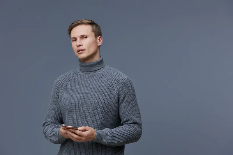 A young man with fair skin and short light brown hair wearing a cozy gray knit sweater holding a smartphone in his hands, looking thoughtfully at the camera against a plain grey background. People lif