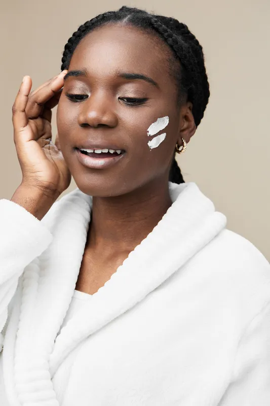 Young Black woman applying face cream, showing joy and self care with a minimalistic beige background for skincare concepts